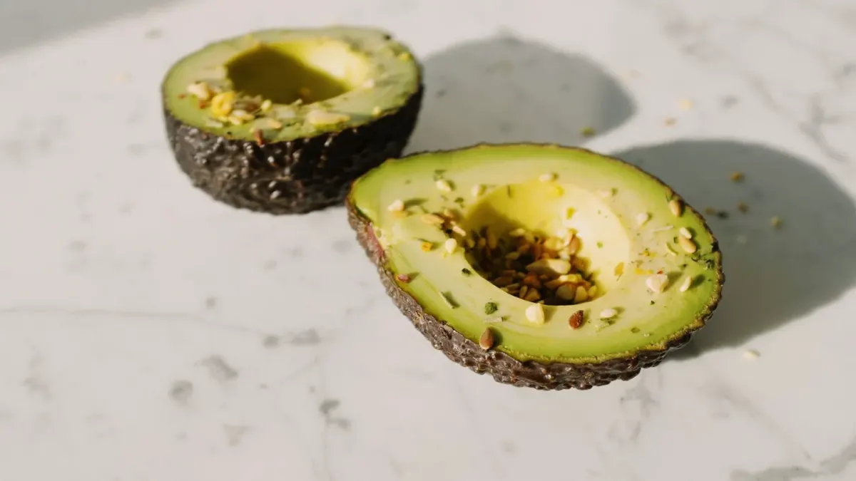 A halved avocado on a cutting board with a dog sniffing nearby on a kitchen floor